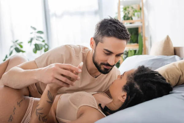Homme barbu souriant tenant la main de femme afro-américaine tatouée en lingerie sexy allongée sur le lit à la maison — Photo de stock