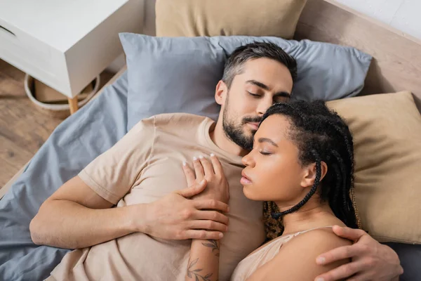 Vista superior do homem barbudo com olhos fechados abraçando mulher americana africana sensual enquanto deitado na cama em casa — Fotografia de Stock