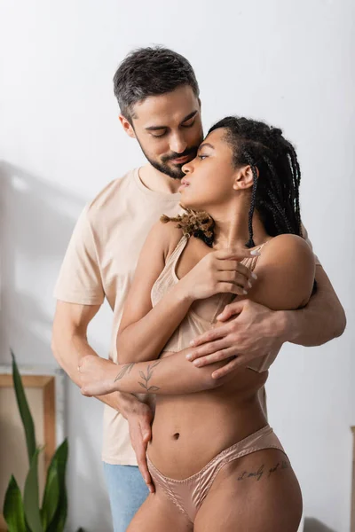 Татуированная африканская американка в сексуальном белье рядом с бородатым мужчиной в футболке в спальне дома — стоковое фото