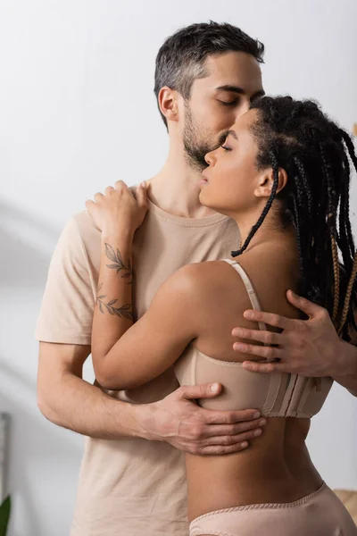 Bruna uomo in t-shirt che abbraccia tatuata donna africana americana in biancheria intima sexy in camera da letto a casa — Foto stock