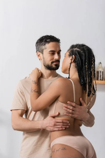 Bearded man in t-shirt hugging seductive african american woman in underwear in bedroom at home — Stock Photo