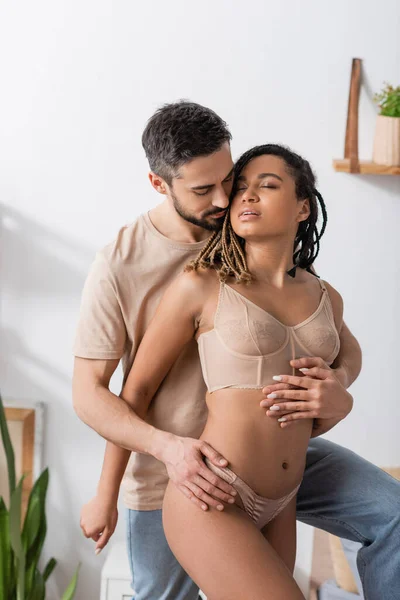 Morena hombre en camiseta y jeans abrazando caliente africana americana mujer en ropa interior sexy en el dormitorio en casa - foto de stock