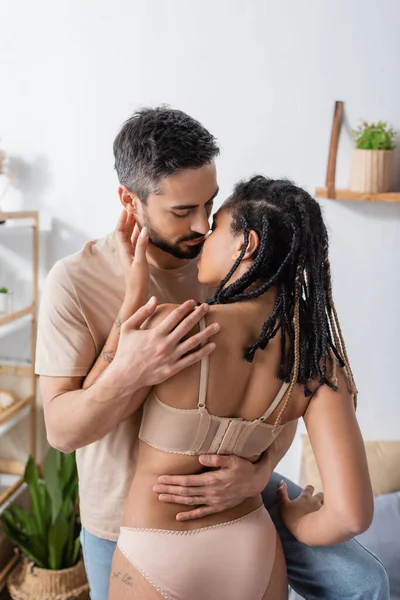 Bruna uomo in t-shirt baciare appassionata donna africana americana con dreadlocks in camera da letto a casa — Foto stock