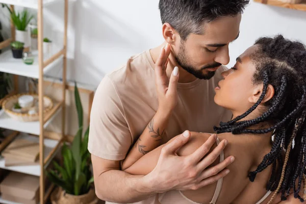 Bearded brunette man in t-shirt hugging seductive african american woman with dreadlocks in bedroom at home — Stock Photo