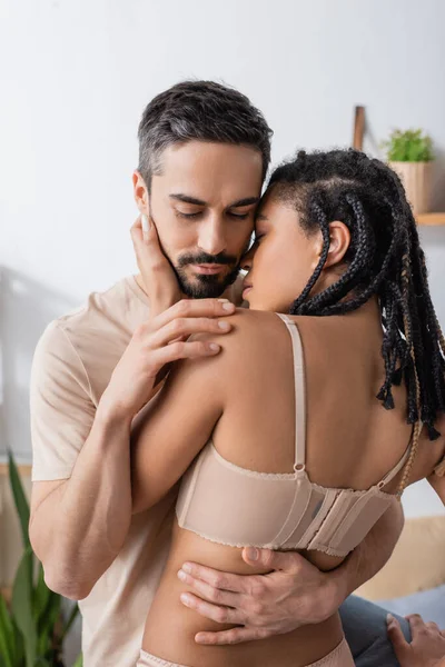Barbudo hombre abrazando sexy africana americana mujer en ropa interior en dormitorio en casa — Stock Photo