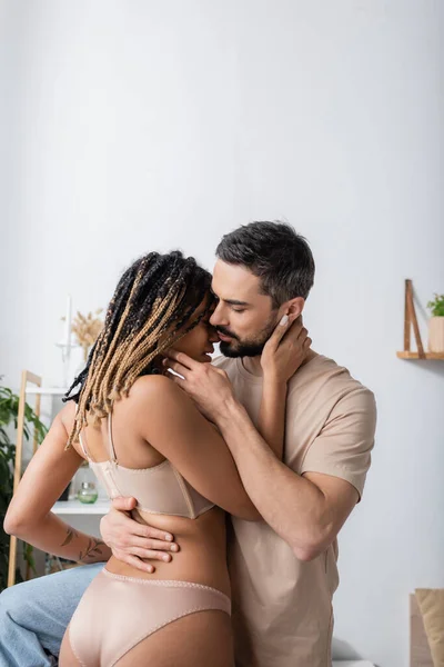 Apasionada mujer afroamericana en lencería besándose con barbudo hombre en camiseta en el dormitorio en casa - foto de stock