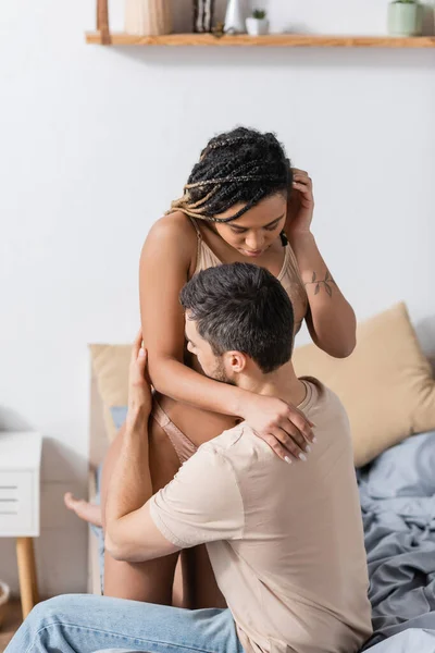 Sexy africaine américaine femme avec dreadlocks séduire brunette homme en t-shirt assis sur le lit à la maison — Photo de stock