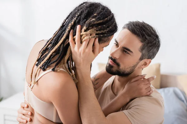 Brunette bearded man embracing sexy african american woman with dreadlocks in bedroom at home — Stock Photo