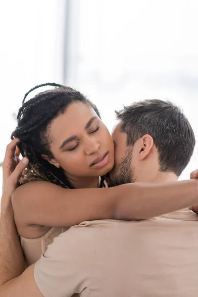 Sexy africano americano mujer con cerrado ojos abrazando morena hombre en casa - foto de stock