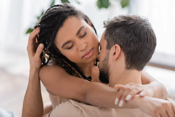 Junge und sexy afrikanisch-amerikanische Frau mit geschlossenen Augen und Dreadlocks umarmt brünetten Mann zu Hause — Stockfoto