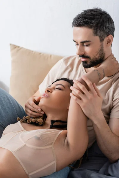 Tattooed african american woman in sexy bra lying with closed eyes near bearded man on bed at home — Stock Photo