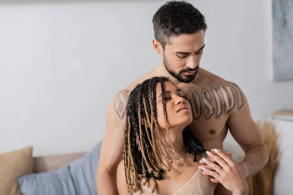 Без сорочки татуйований чоловік спокушає молоду і пристрасну афроамериканську жінку з дредлоками в спальні вдома — стокове фото