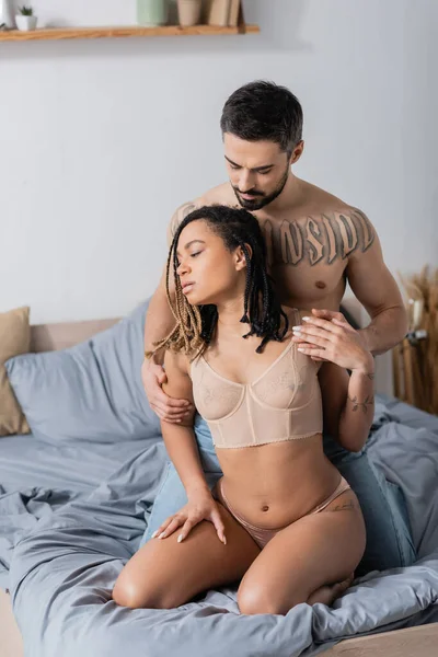 Без рубашки татуированный мужчина соблазняет чувственную африканскую американку в сексуальном белье на кровати дома — стоковое фото