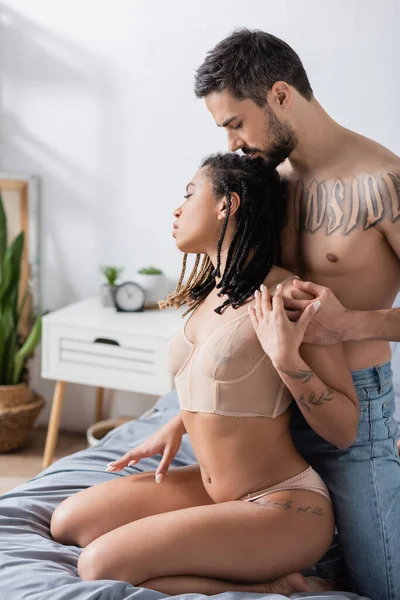 Side view of shirtless tattooed man hugging young african american woman in sexy lingerie in bedroom at home — Stock Photo