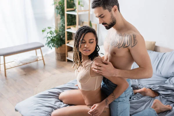 Donna barbuta tatuata che abbraccia giovane e sexy donna africana americana in biancheria intima in camera da letto moderna — Foto stock
