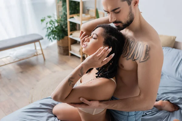 Side view of shirtless tattooed man embracing sexy african american woman in underwear in bedroom at home — Stock Photo