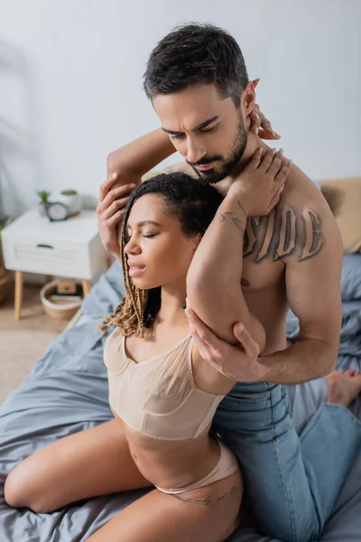 Giovane e calda donna africana americana in reggiseno abbracciando barbuto uomo senza maglietta in camera da letto a casa — Foto stock