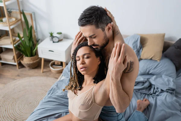 Соблазнительная африканская американка с закрытыми глазами рядом с бородатым мужчиной в спальне дома — стоковое фото