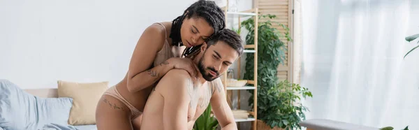 Татуйована і сексуальна афроамериканська жінка обіймає бородатого без сорочки чоловіка в спальні вдома, банер — стокове фото