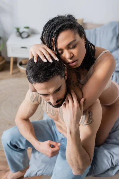 Mujer afroamericana sensual con los ojos cerrados abrazando sin camisa hombre tatuado sentado en la cama en casa - foto de stock