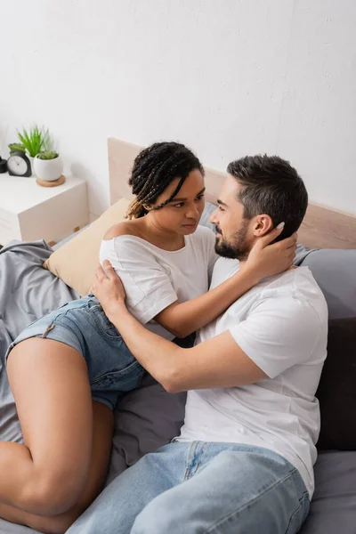 High angle view of multiracial couple in white t-shirts embracing and looking at each other on bed at home — Stock Photo