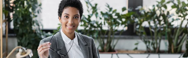 Successful african american businesswoman holding eyeglasses and smiling at camera in office, banner — Stock Photo