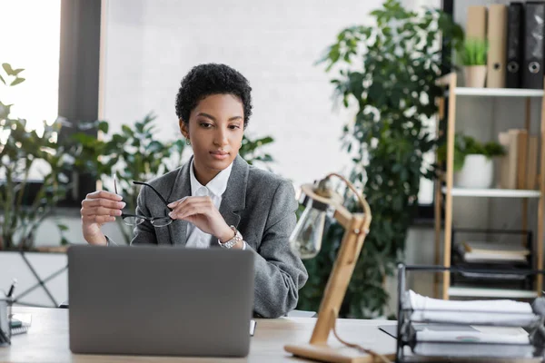Élégant gestionnaire afro-américain tenant des lunettes tout en étant assis près d'un ordinateur portable et lampe de bureau floue dans le bureau — Photo de stock
