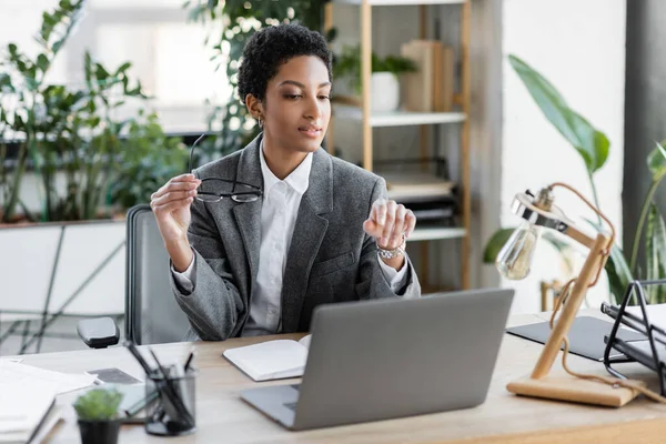 Africano americana empresária em blazer cinza segurando óculos e apontando para laptop no escritório moderno — Fotografia de Stock