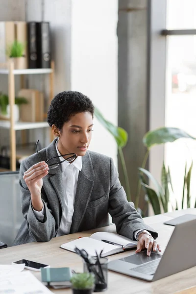 Афроамериканська бізнесменка в сірому блейзері тримає окуляри і використовує ноутбук в сучасному офісі. — Stock Photo