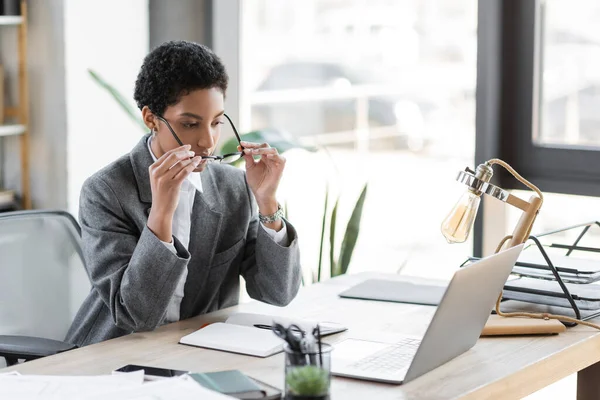 Mulher de negócios americana africana elegante colocando óculos enquanto sentado perto de laptop e notebook em branco no escritório — Fotografia de Stock
