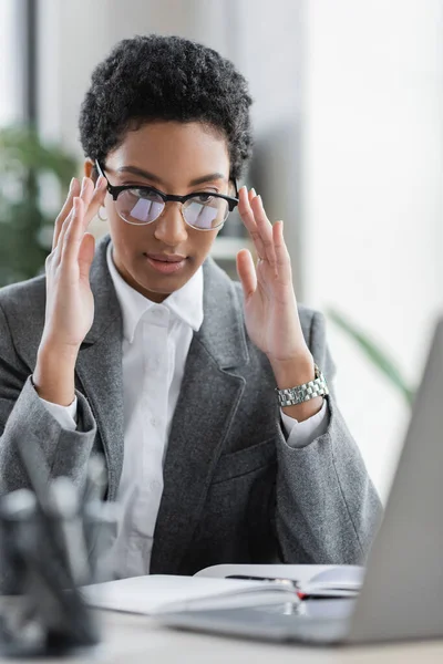 Giovane donna d'affari africana americana in giacca grigia che regola gli occhiali da vista alla moda vicino al computer portatile offuscato in ufficio — Foto stock