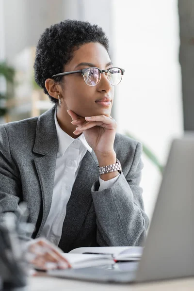 Вдумчивая американская бизнесвумен в сером пиджаке и очках, смотрящая в сторону в офисе — стоковое фото