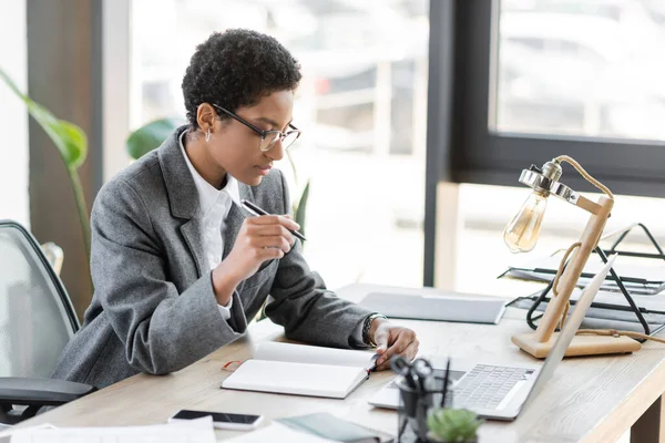 Stylish african american businesswoman in grey blazer and eyeglasses sitting with pen near blank notebook and laptop in modern office — Stock Photo