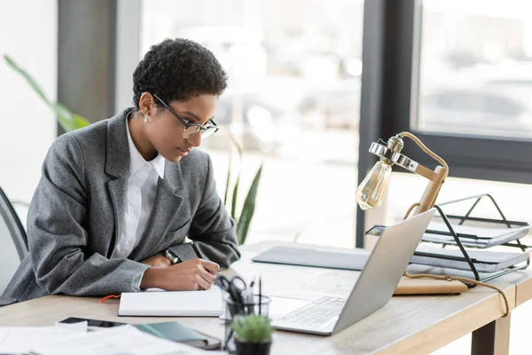 Junge afrikanisch-amerikanische Geschäftsfrau mit Brille schaut auf Laptop und schreibt in Notizbuch, während sie im Büro arbeitet — Stockfoto