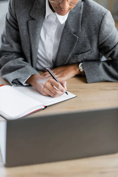 Cropped view of african american businesswoman in grey blazer writing in blank notebook on desk in office — Stock Photo