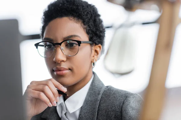 Portrait of african american businesswoman in grey blazer and eyeglasses holding hand near face while thinking in office — Stock Photo