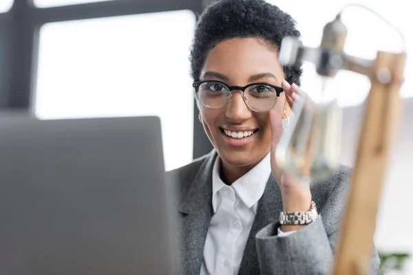 Retrato de mulher de negócios americana africana alegre ajustando óculos elegantes e olhando para a câmera perto de laptop borrado no escritório — Fotografia de Stock