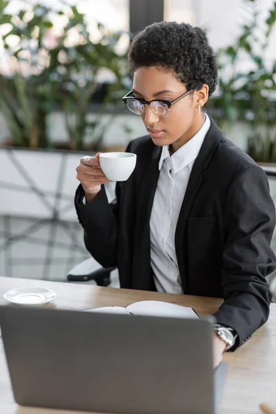Élégante femme d'affaires afro-américaine en blazer noir et lunettes assis avec tasse de café près d'un ordinateur portable flou au bureau — Photo de stock