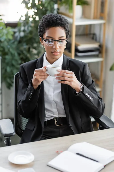 Afro-americana donna d'affari in elegante abbigliamento formale e occhiali da vista in possesso di tazza di caffè e guardando notebook in bianco in ufficio — Foto stock
