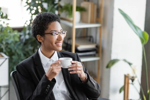 Mulher de negócios americana africana alegre em blazer preto e óculos segurando xícara de café e olhando para longe no escritório — Fotografia de Stock