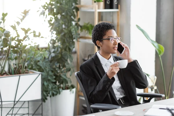 Young african american businesswoman with coffee cup talking on mobile phone in modern office — Stock Photo