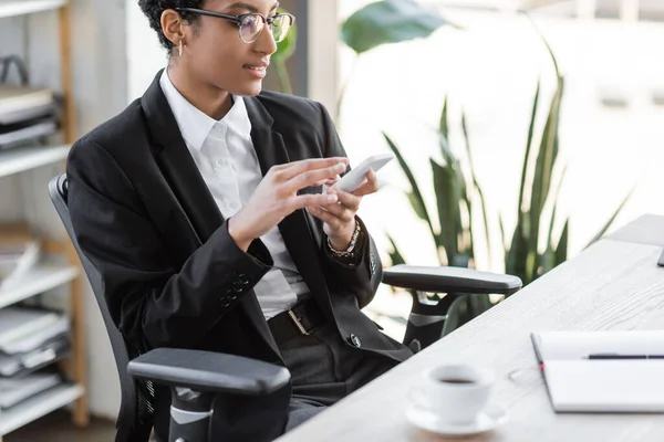 Positive african american businesswoman in eyeglasses and stylish blazer messaging on mobile phone near blurred coffee cup in office — Stock Photo