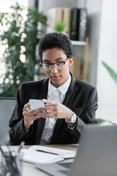 African american businesswoman in stylish formal wear and eyeglasses messaging on smartphone in office — Stock Photo