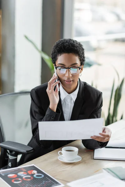 African american businesswoman in eyeglasses and black blazer looking at document and calling on smartphone in office — Stock Photo