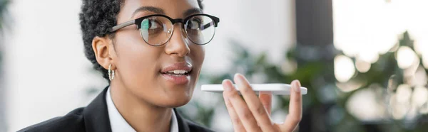 Portrait d'une femme d'affaires afro-américaine aux lunettes élégantes envoyant un message vocal sur smartphone au bureau, bannière — Photo de stock