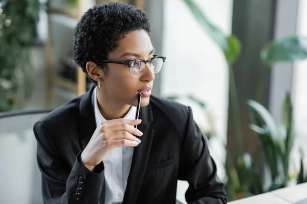 Pensativa mujer de negocios afroamericana en gafas y blazer negro sosteniendo pluma y mirando hacia otro lado en la oficina - foto de stock