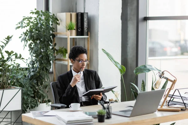 Розсудлива афроамериканська бізнесменка, що тримає ручку і дивиться на записник біля ноутбука і документи в сучасному офісі — стокове фото