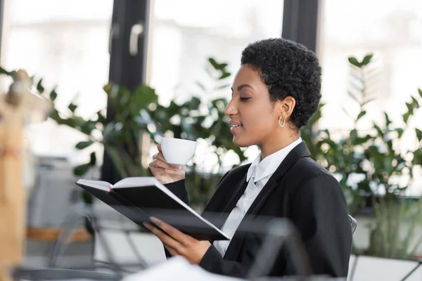 Vista lateral da empresária afro-americana sentada com caderno e xícara de café no escritório — Fotografia de Stock