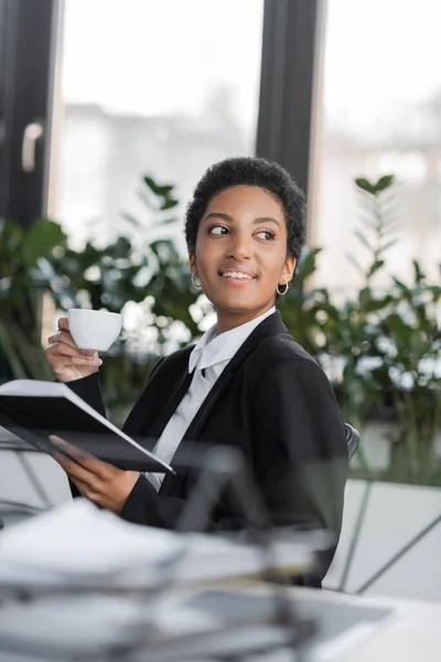 Gioiosa donna d'affari afro-americana che distoglie lo sguardo mentre si siede con tazza di caffè e notebook in ufficio — Foto stock