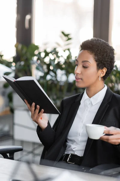 Elegante mujer de negocios afroamericana con taza de café mirando el cuaderno mientras está sentado en la oficina - foto de stock